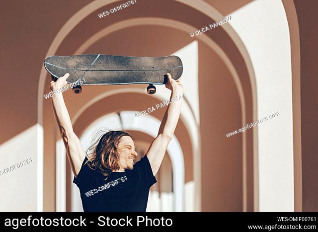 Man with long hair lifting skateboard above head in arcade