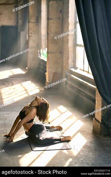 Delightful girl in a dark sportswear engaged in yoga on the windows background in a loft style hall. She does a half split on the mat and leans on her hands