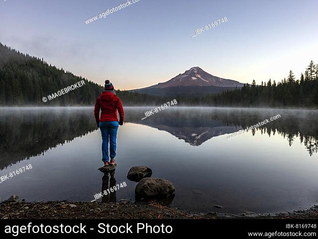 Young woman standing on a stone, reflection of Mt. Hood volcano in Trillium Lake, at sunrise, Oregon, USA, North America