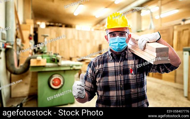 Carpenter worker at work in the carpentry workshop wears surgical mask to prevent Coronavirus spread, makes OK sign with thumb up
