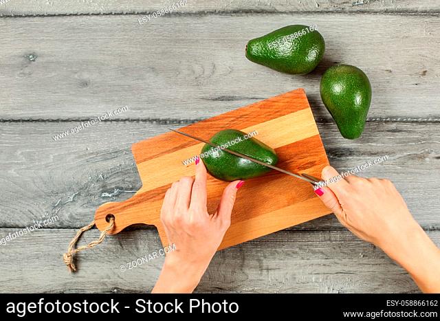 Tabletop view, woman hands holding chef knife, cutting avocado on chopping board, two whole avocados near on gray wood desk