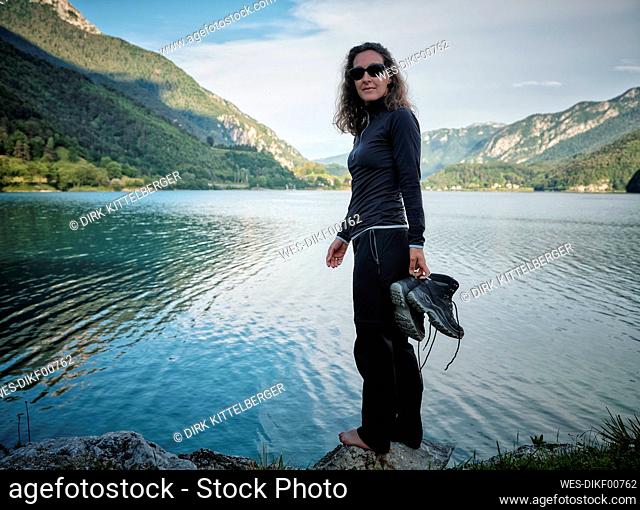 Smiling woman holding boots standing on rock at lake