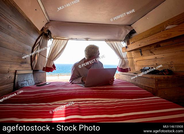 inside view of a minivan with a young blonde man using his laptop and working near of the beach and the sea - traveler and nomad people lifestyle having fun