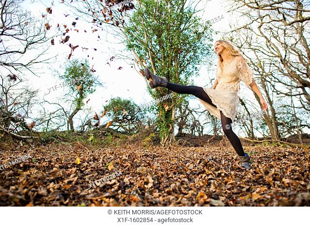 a slim blonde woman girl alone in woodland autumn afternoon daytime kicking up dry leaves, UK