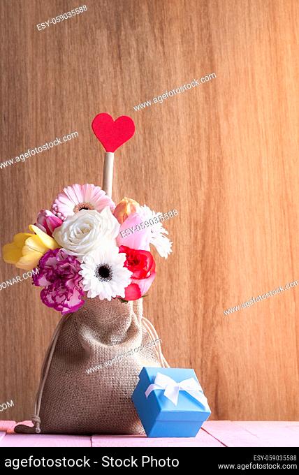 Valentine day card with a beautiful bouquet of flowers in a jute bag, with a red heart and a blue gift box, on a pink table and a wooden background