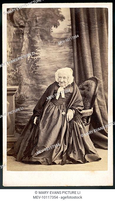 An old woman is photographed seated in an upholstered chair. Her clothing is in dark, sombre colours & she wears a lace bonnet & shawl