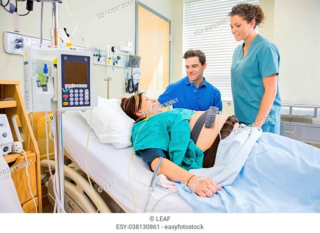 Happy nurse and man looking at pregnant woman lying on bed in hospital