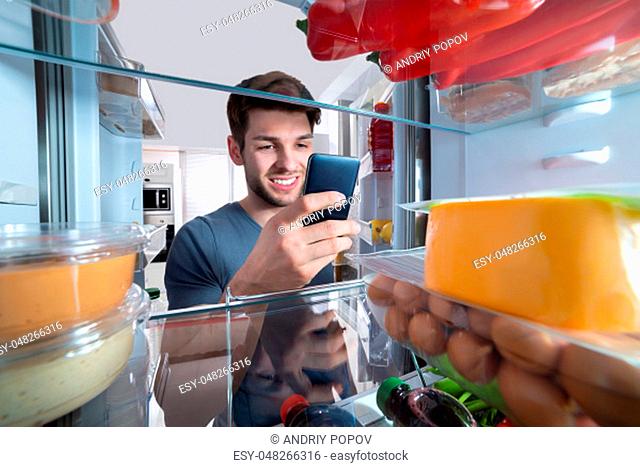 Happy Man Standing In Front Of Refrigerator Using Mobilephone In Kitchen
