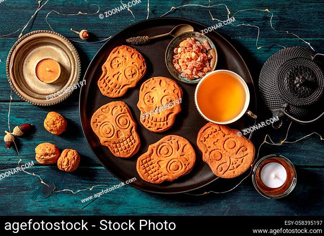 Halloween ginger cookies in the shape of skulls, homemade Dia de los muertos biscuits, shot from above with tea and candles on a dark wooden background