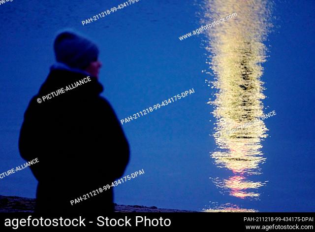 18 December 2021, Saxony-Anhalt, Muldestausee: A walker stands at the Goitzsche Lake while the illuminated water level tower is reflected in the water