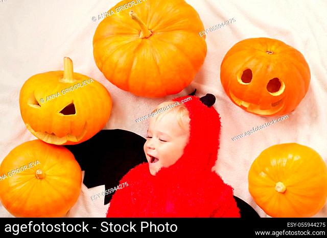Baby in devil costume with pumpkins. Child in halloween outfit lying on soft white blanket