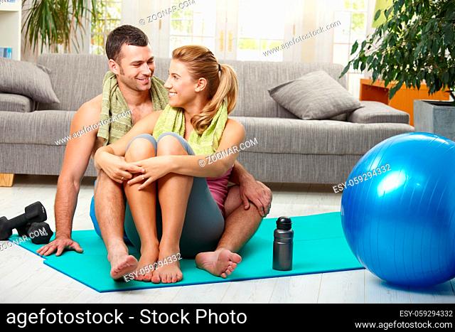 Happy couple resting after training, sitting on fitness mat in living room, looking at each other smiling