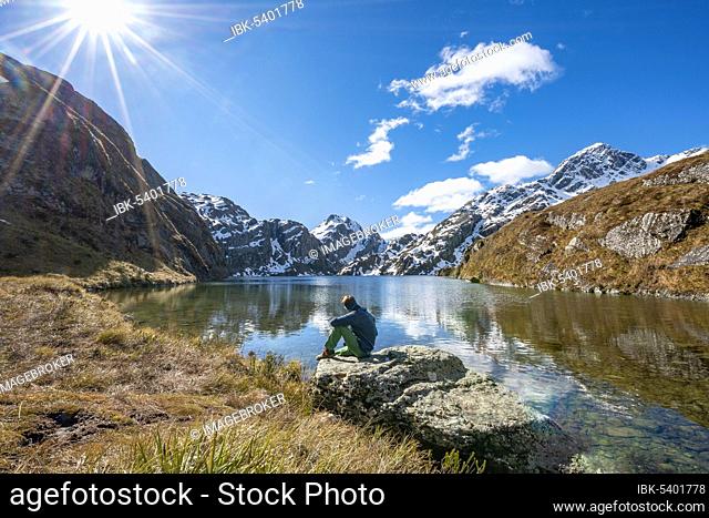 Young man sitting on a rock, mountains reflected in the lake, Lake Harris, Conical Hill, Routeburn Track, Mount Aspiring National Park, Westland District