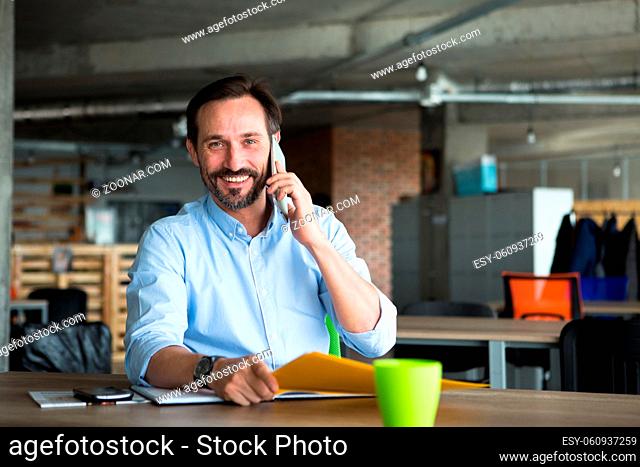 Handsome businessman talking over mobile or smart phone and looking at camera while communicating with foreign business partners in office interior