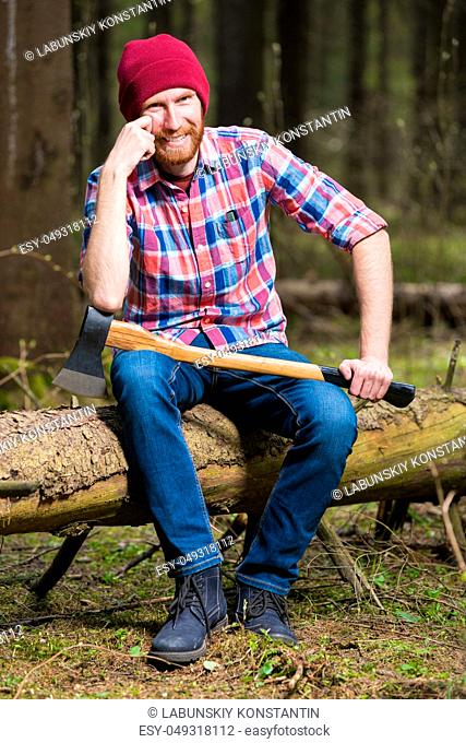 happy bearded lumberjack with an ax is sitting on a log in the forest
