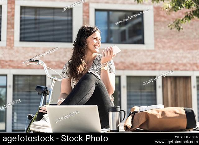 Smiling woman talking over smart phone while sitting against building in city