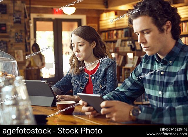 Young man and woman sitting at counter in cafe bookstore using digital tablets