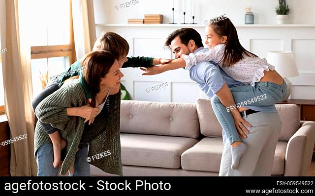 Happy young married couple giving piggyback ride to little kids siblings, playing funny game together at home. Overjoyed parents holding on back small children...