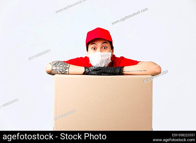 Excited asian delivery guy in medical mask and protective gloves, leaning on box parcel and staring camera astonished. Courier working during coronavirus...