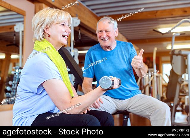 Senior woman in the gym exercising with dumbbells for fitness while her husband fires her up