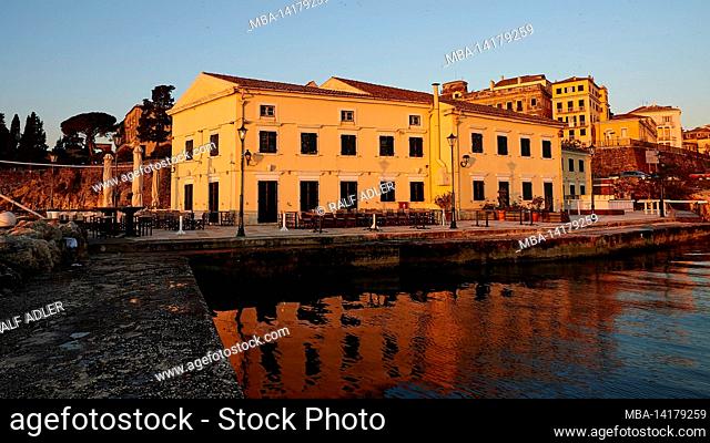Greece, Greek Islands, Ionian Islands, Corfu, Corfu Town, sunrise by the sea, buildings by the sea in the morning light, cafe