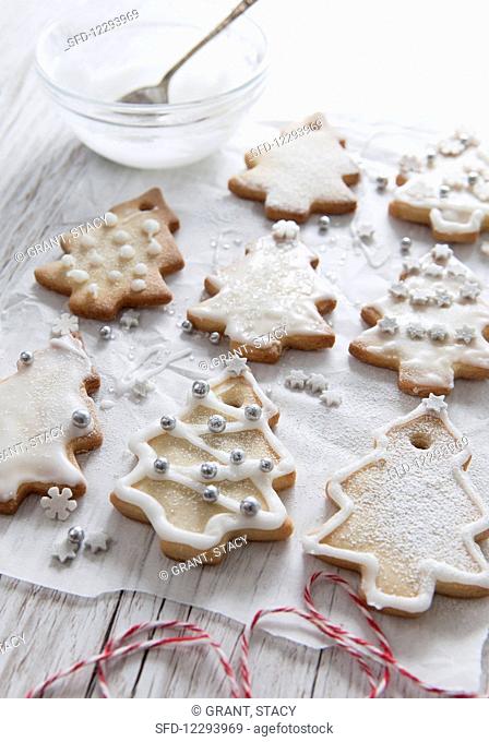 Tree shaped Christmas biscuits being decorated with white icing and christmas sprinkles bakers twine in the forground and icing bowl and silver sppon in the...