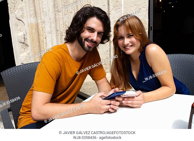 Young couple looking at mobile on a summer terrace, smile looking at camera