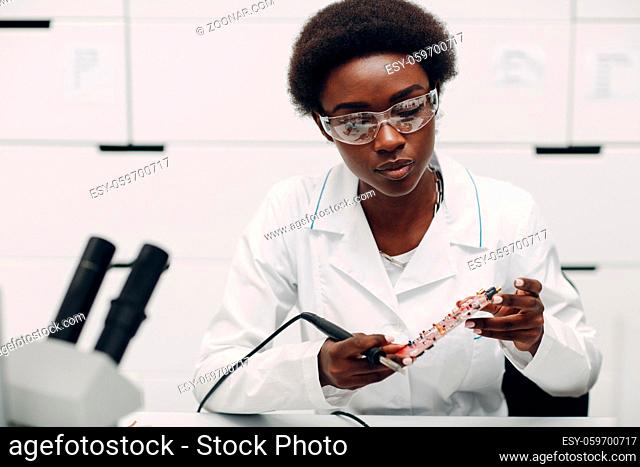 Scientist african american woman working in laboratory with soldering ironand checking electronic plate board. Research and development of electronic devices by...
