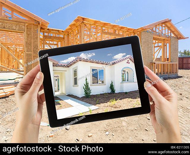 Female hands holding computer tablet with finished house on screen, construction framing behind