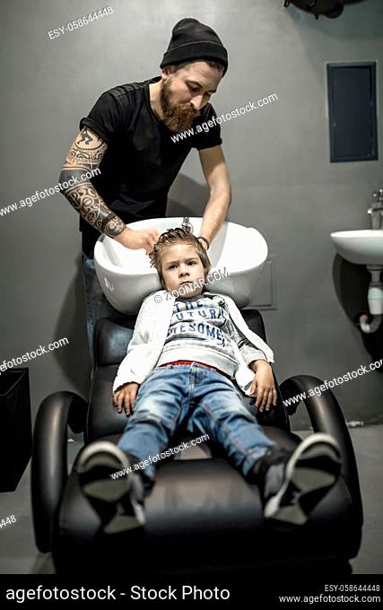 Little boy in blue jeans in the barbershop. He lies on the armchair and bearded tattooed barber washes his hair in the white sink
