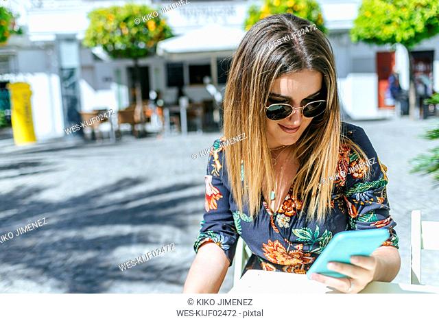 Spain, Cadiz, Vejer de la Frontera, young woman sitting at street cafe looking at mobile phone