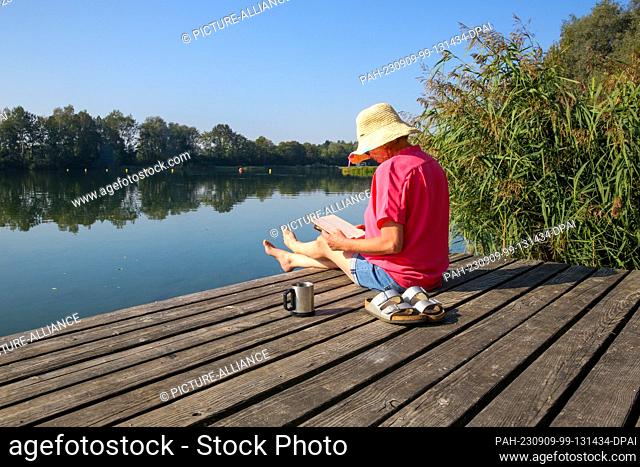 09 September 2023, Baden-Württemberg, Ertingen: A woman sits on a bathing platform in the sun with a book in her hand and a cup of coffee