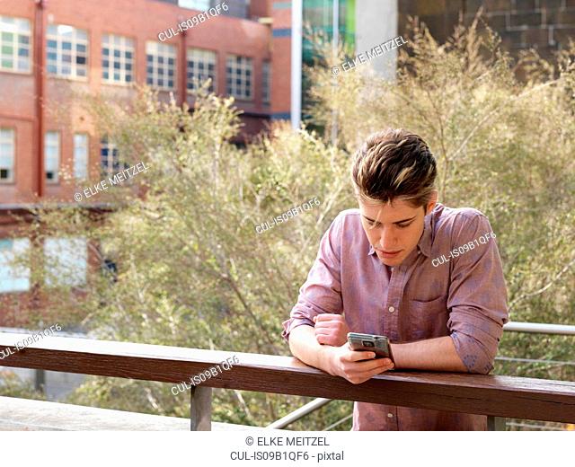 Young man leaning on fence, using smartphone, outdoors
