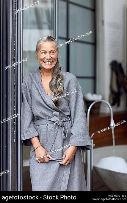 Smiling mature woman wearing bathrobe leaning by glass door in bathroom