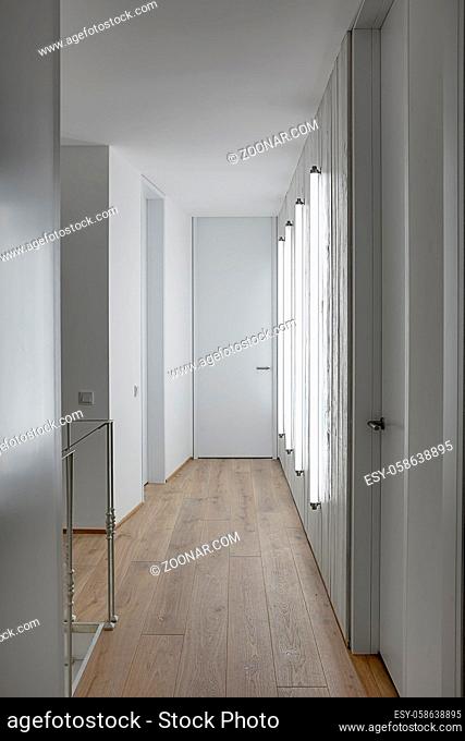 Light corridor in a modern style with white walls and a parquet on the floor. There are glowing tube lamps on the wooden wall, doors, metal railing, switches