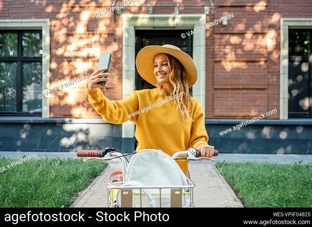 Smiling woman with bicycle taking selfie through smart phone in front of building