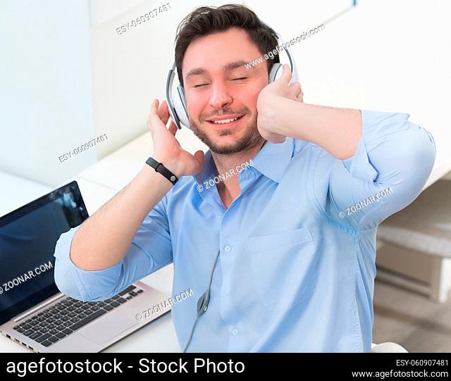 Portrait of handsome and happy man with earphones on listening to music while working on laptop computer