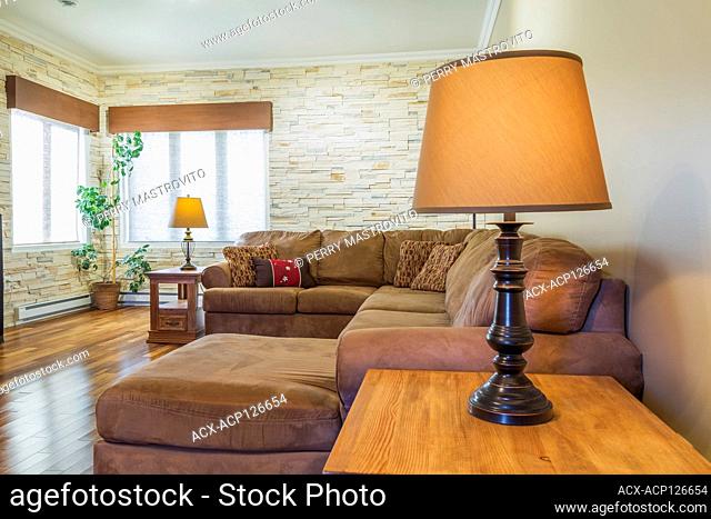Tan sectional sofas and table lamp in upstairs family room with imitation stone wall inside a country cottage style house, Quebec, Canada