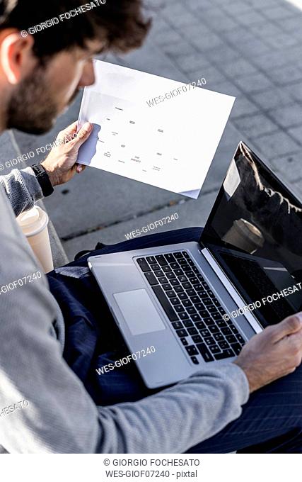 Man sitting outdoor using laptop and reviewing paper
