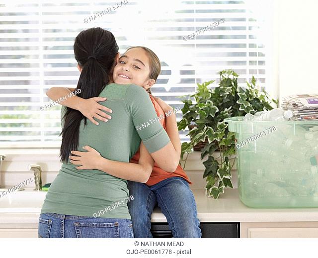 Woman and young girl in kitchen hugging by bin of recyclable materials