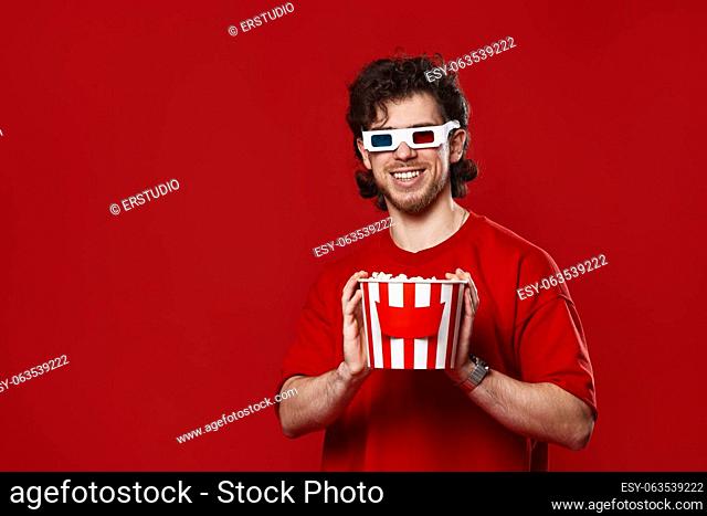 handsome bearded man in 3d glasses holding big bucket of popcorns on red background