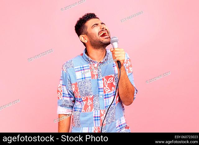 Excited positive man with beard in casual blue shirt singing songs, holding microphone, singer making performance, keeps eyes closed