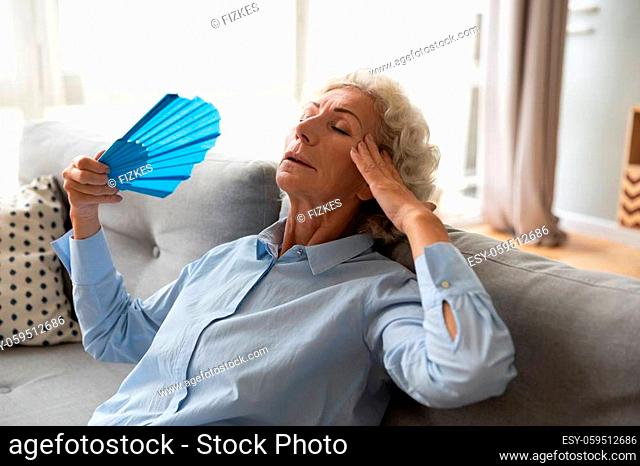Stressed overheated elderly female leaned on couch holds blue colour fan relieving high temperature refreshing herself in living room without air-conditioner...