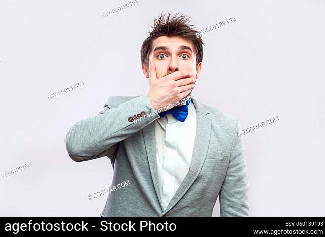 Portrait of shocked handsome bearded man in casual grey suit and blue bow tie standing covering his mouth and looking at camera with big eyes