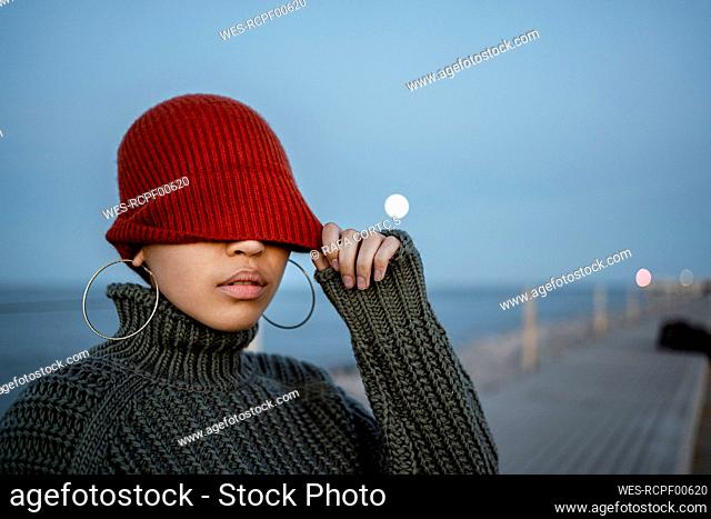 Woman covering eyes with knit hat while standing against sky