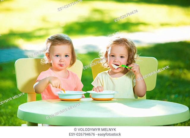 Two little 2 years old girls sitting at a table and eating together against a green lawn