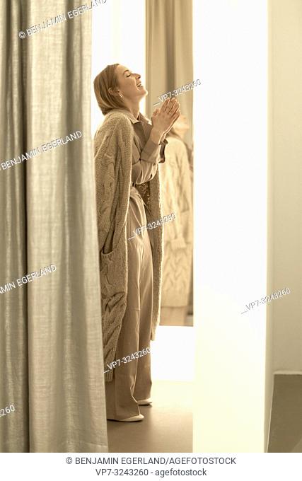 happy fashionable woman with folded hands inside fitting room of clothing store, wearing new stylish one-piece jumpsuit combined with beige coat