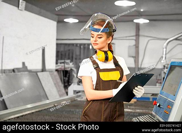 Front view of smiling female worker of metal factory in process of writing data. Pretty woman in uniform and protective mask standing near computer and cnc and...