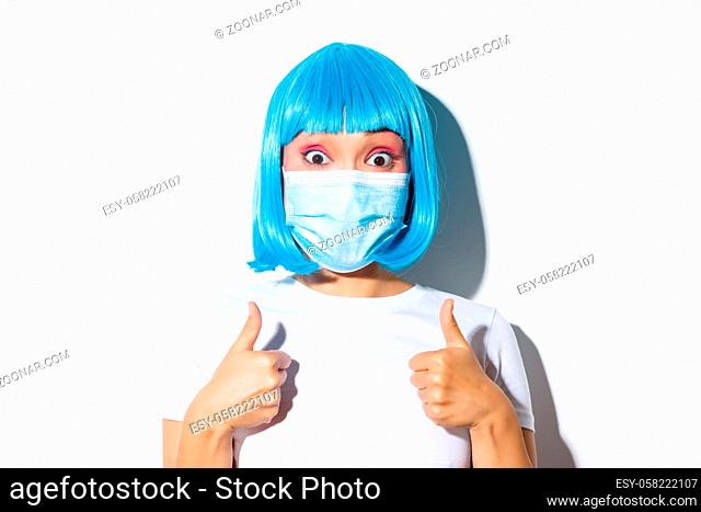 Concept of halloween celebration and coronavirus. Close-up of excited asian girl in medical mask and blue wig showing thumbs-up and approve something good