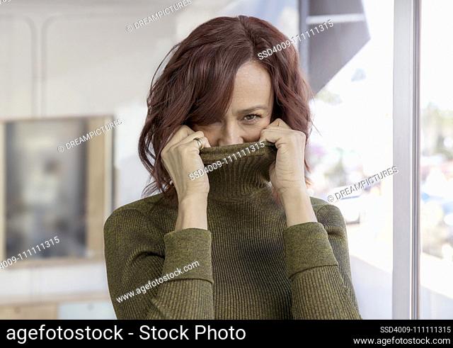 A playful middle-aged woman hides her face in her turtleneck sweater